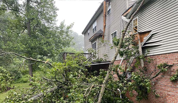A house with buckled siding and debris from a fallen tree after a severe storm